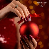 Hand with red glitter nails holding a red Christmas ornament, highlighting SPN Nails UV/LED Gel Polish 1023 Kiss the Star.